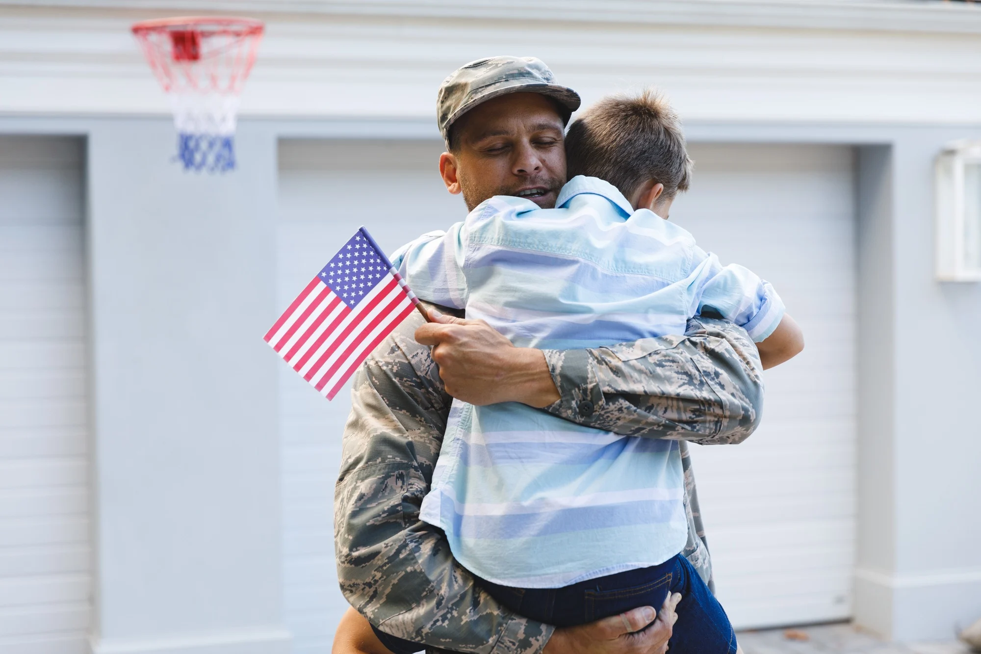 Smiling caucasian male soldier with son outside house holding american flags